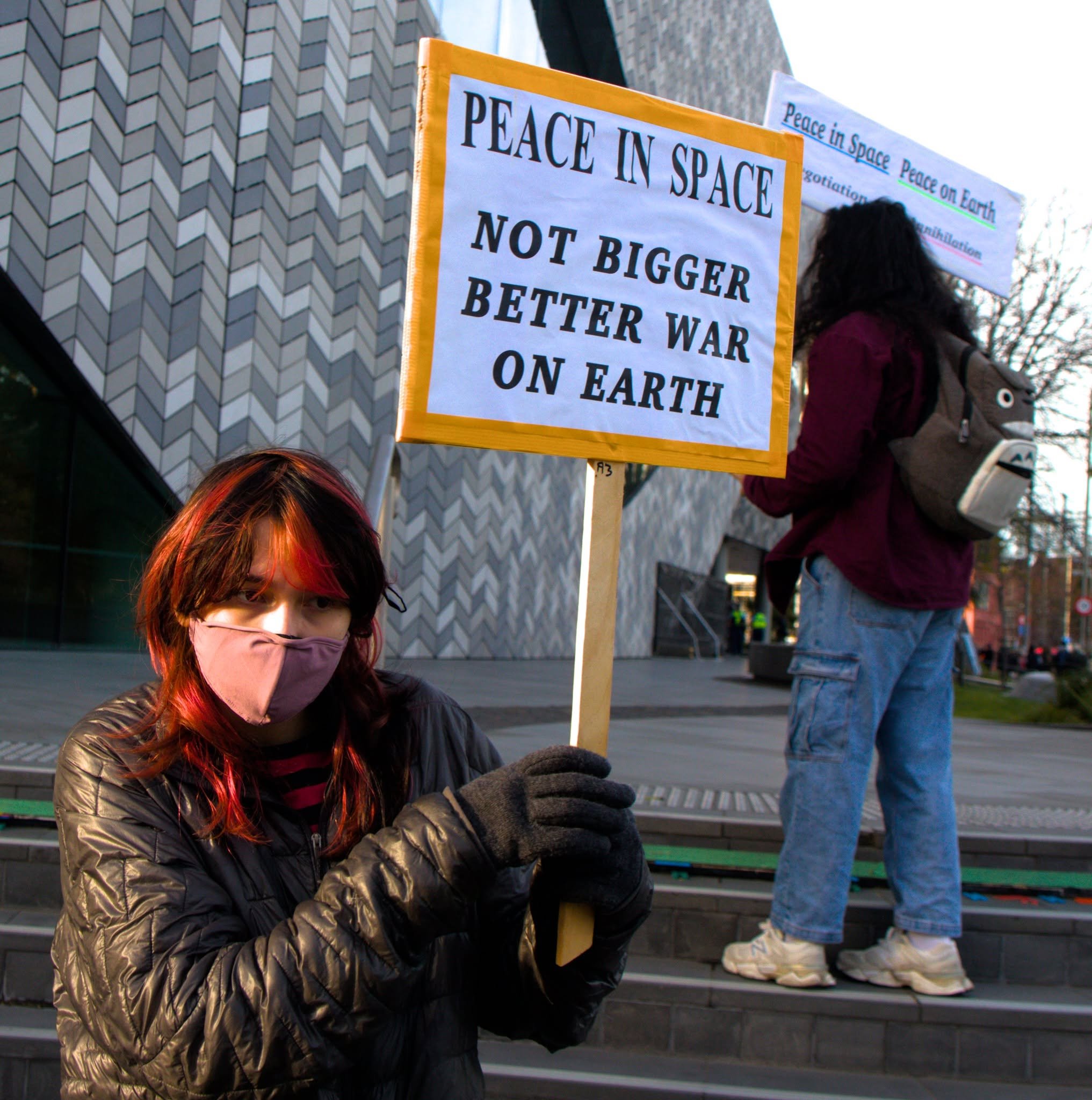 A protester holding a sign reading 'Peace in Space: Not Bigger Better War on Earth' outside Te Pae Convention Centre